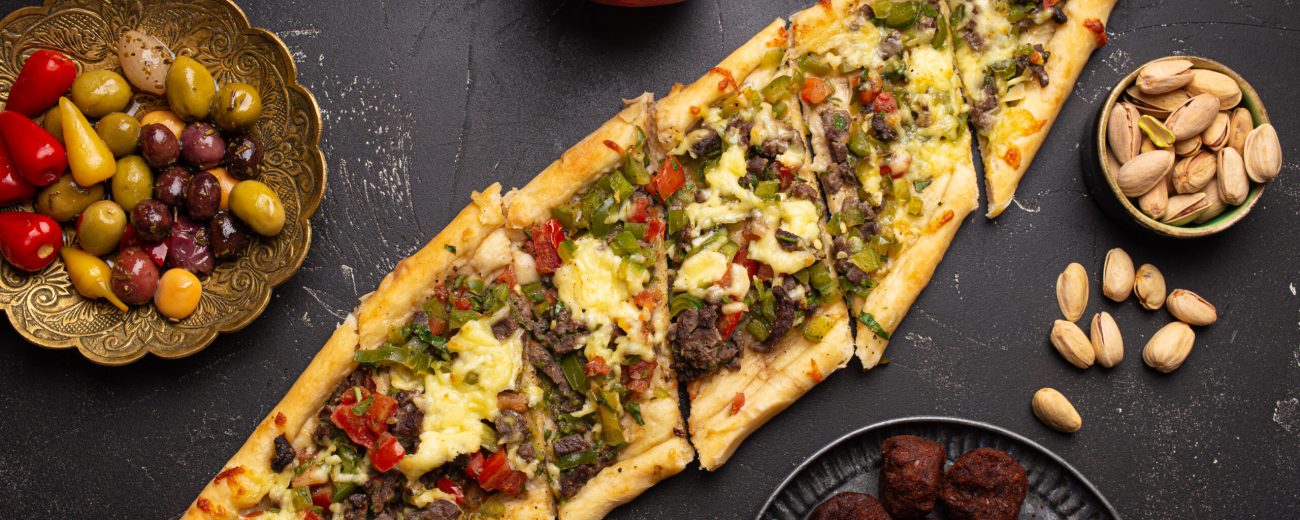 Traditional Turkish pizza pide freshly baked with ground beef and vegetables served on black stone background with assorted Middle Eastern meze from above, cuisine of Turkey